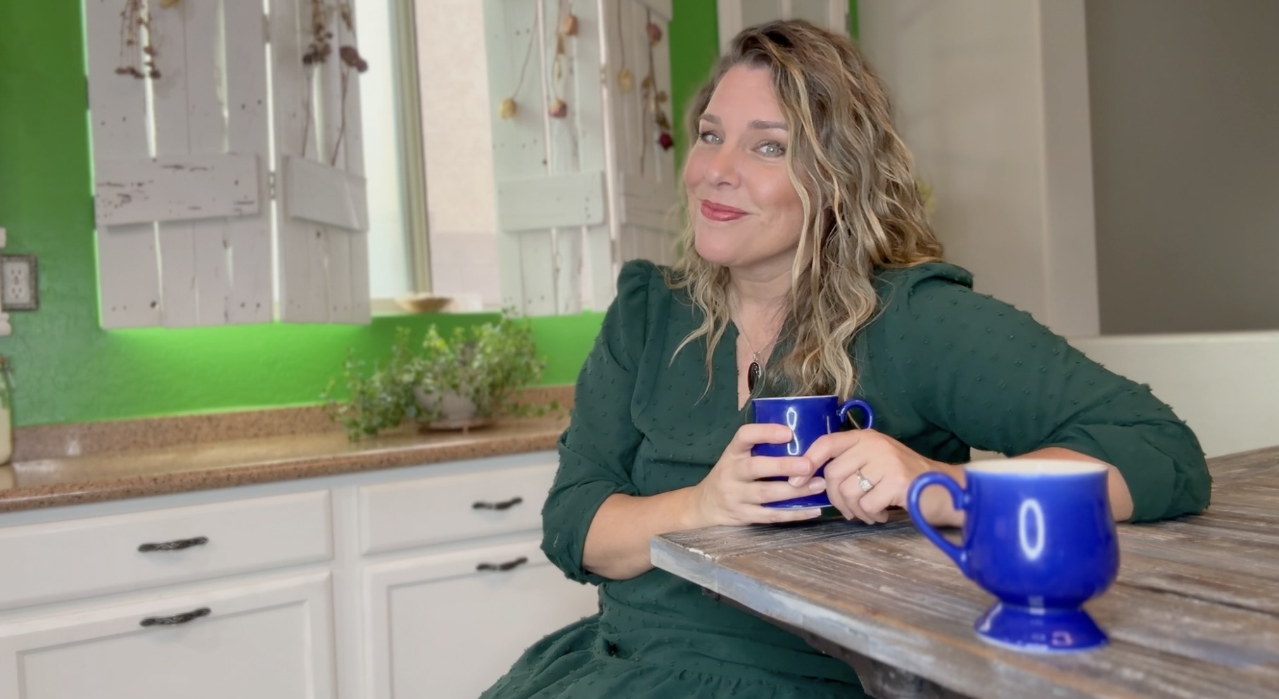 Heather smiling at a rustic wooden table, holding a blue mug, with a second blue mug set out in front of the viewer