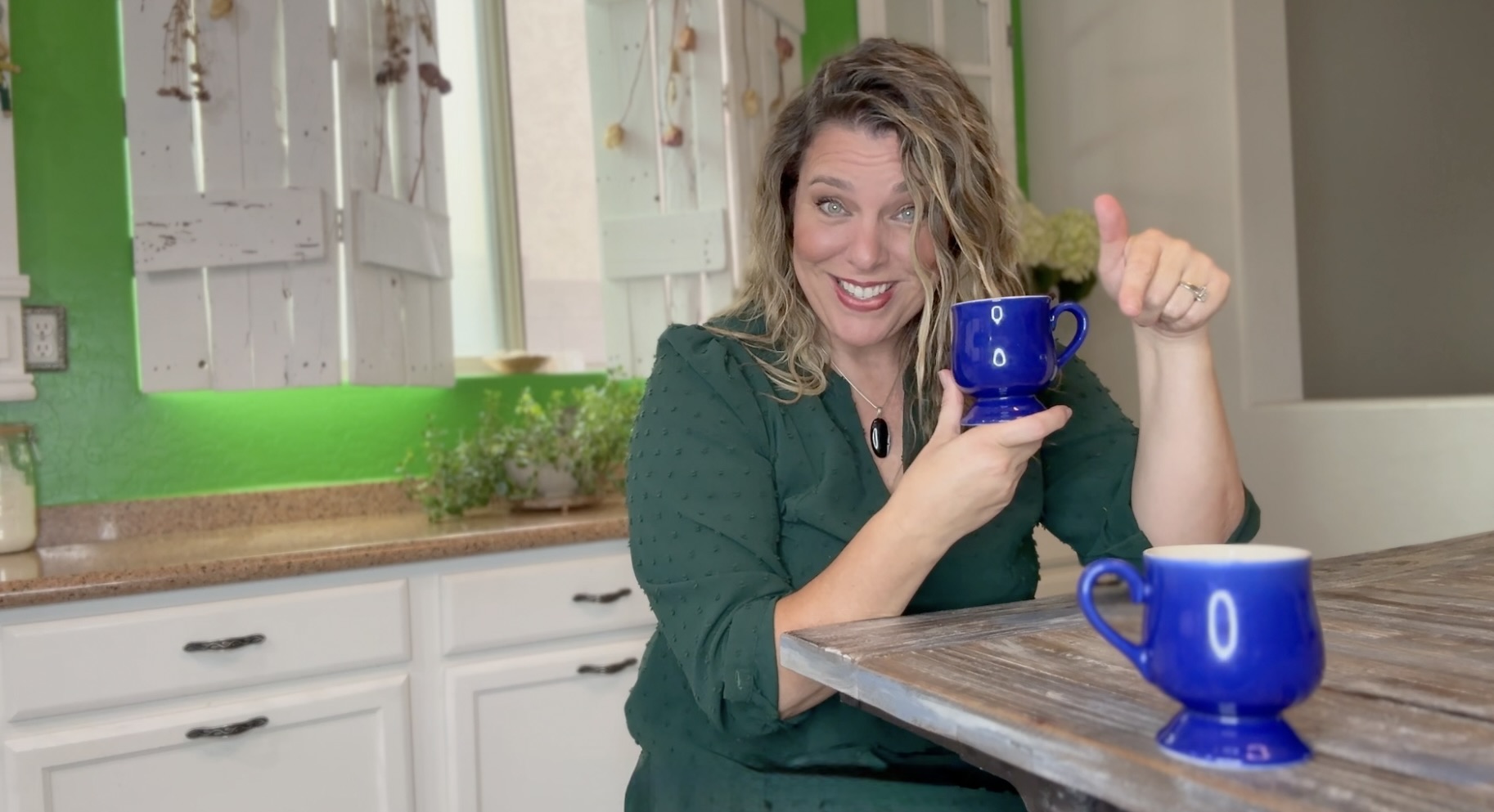 Heather sitting at a rustic table, holding a mug, pointing at a second mug